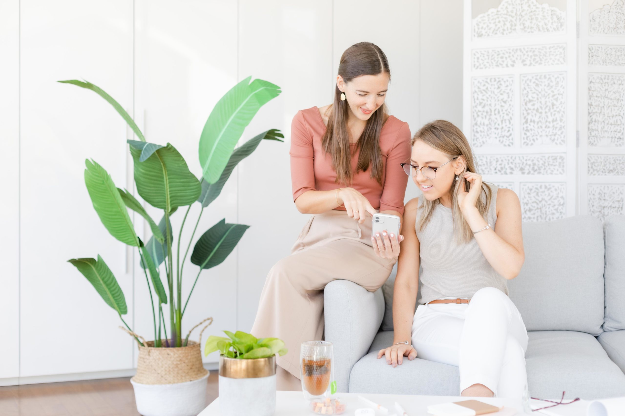 two girls sitting on white couch. One on left holding phone and pointing. Girl on right looking with hand holding hair behind her ear. Green floor plant on the left of the two girls.
