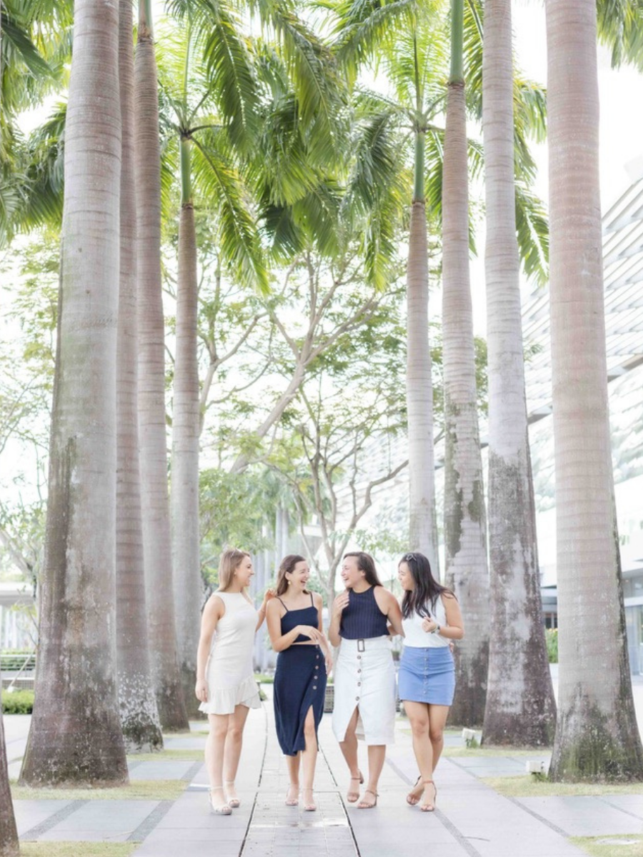 four girls walking toward the camera looking at each other laughing. Tall palm trees on either side of them.