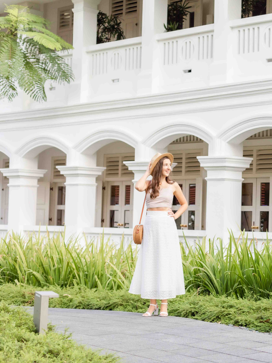 White building in the background with archways. Tall green grass in front of the building with a palm tree in the upper left corner. Krystle dressed in white standing in front of the grass with right hand on her hip looking to the right.