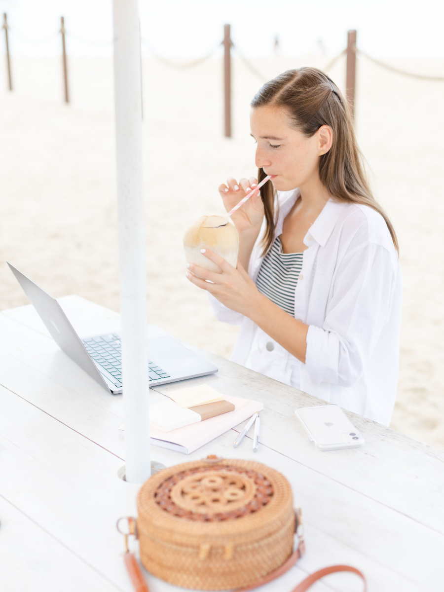 Krystle sitting at a white picnic table on the beach with a computer in front of her, a wicker purse to her left while she is sipping on a drink.