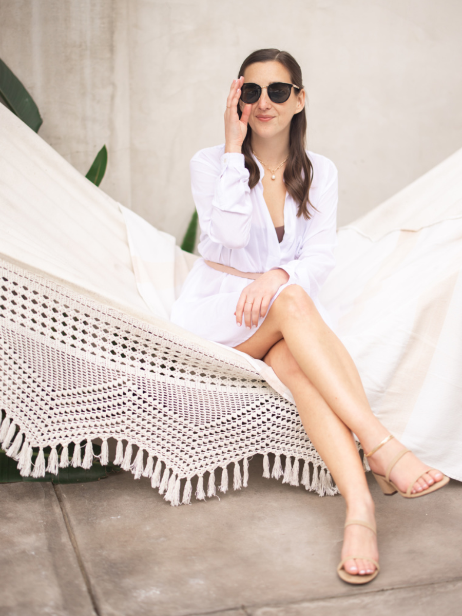 Krystle wearing all white and black sunglasses sitting on a white crochet hammock. Legs crossed. Hand on sunglasses. Soft smile.