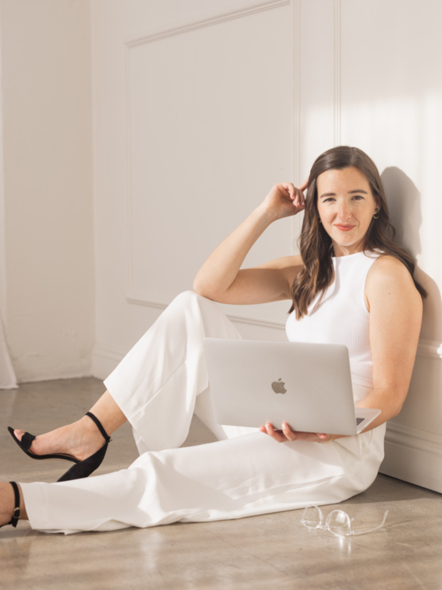 Krystle sitting on the floor. Leg closest to camera is straight out with the other leg behind it bend with her elbow resting on it. In her other hand she is holding up a silver macbook laptop while soft smiling toward the camera.